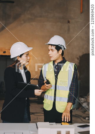 Construction manager and engineer dressed in orange work vests and hard helmets explore construction documentation on the building site near the steel frames 128969420