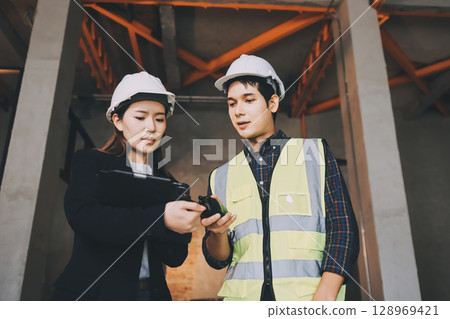Construction manager and engineer dressed in orange work vests and hard helmets explore construction documentation on the building site near the steel frames 128969421