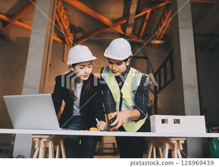 Construction manager and engineer dressed in orange work vests and hard helmets explore construction documentation on the building site near the steel frames Construction manager and engineer dressed in orange work vests and hard helmets explore construction documentation on the building site near the steel frames 128969439
