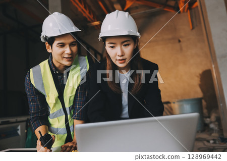 Construction manager and engineer dressed in orange work vests and hard helmets explore construction documentation on the building site near the steel frames Construction manager and engineer dressed in orange work vests and hard helmets explore construction documentation on the building site near the steel frames 128969442