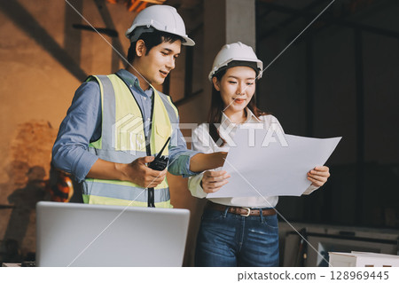 Construction manager and engineer dressed in orange work vests and hard helmets explore construction documentation on the building site near the steel frames Construction manager and engineer dressed in orange work vests and hard helmets explore construction documentation on the building site near the steel frames 128969445