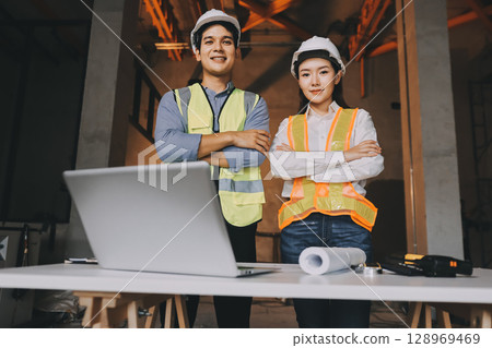 Construction manager and engineer dressed in orange work vests and hard helmets explore construction documentation on the building site near the steel frames Construction manager and engineer dressed in orange work vests and hard helmets explore construction documentation on the building site near the steel frames 128969469