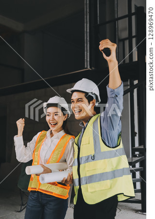 Construction manager and engineer dressed in orange work vests and hard helmets explore construction documentation on the building site near the steel frames 128969470