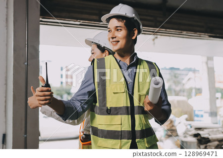 Construction manager and engineer dressed in orange work vests and hard helmets explore construction documentation on the building site near the steel frames Construction manager and engineer dressed in orange work vests and hard helmets explore construction documentation on the building site near the steel frames 128969471