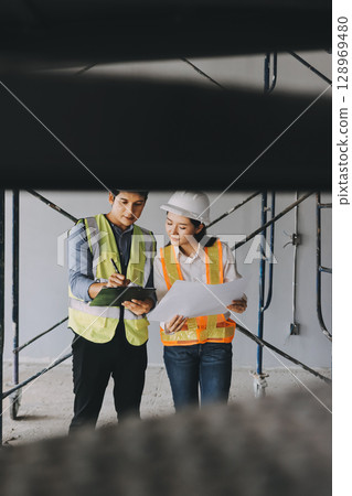 Construction manager and engineer dressed in orange work vests and hard helmets explore construction documentation on the building site near the steel frames 128969480