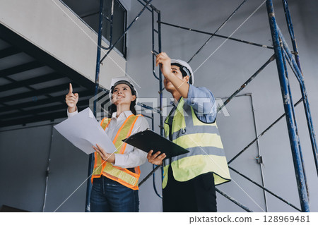 Construction manager and engineer dressed in orange work vests and hard helmets explore construction documentation on the building site near the steel frames Construction manager and engineer dressed in orange work vests and hard helmets explore construction documentation on the building site near the steel frames 128969481