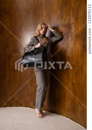 A stylish young woman, long blonde hair, poses in a grey suit, holding a black leather bag against a warm wooden background 128970112