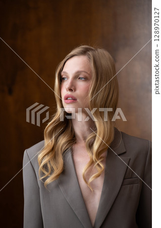 A beautiful young woman with long blonde wavy hair, wearing a stylish grey blazer, gazes thoughtfully to the side against a warm, blurred background A beautiful young woman with long blonde wavy hair, wearing a stylish grey blazer, gazes thoughtfully to the side against a warm, blurred background 128970127