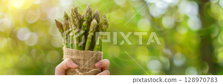 Hand holding a fresh asparagus bouquet wrapped in burlap amid a spring nature background 128970783