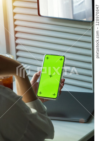 woman hand using hand holding phone mockup with blank green screen with copy space, look at smartphone at home. laptop and ice coffee at background. vertical woman hand using hand holding phone mockup with blank green screen with copy space, look at smartphone at home. laptop and ice coffee at background. vertical 128970847
