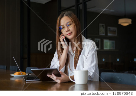 Asian woman wearing eyeglasses talking on the phone,  looking data information at tablet, using laptop computer for work in the morning on the wooden dining table, working from home concept 128970848