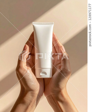 Person Holding White Cream Tube In Hand with Soft Sunlight and Beige Background 128971177