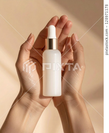Hands Holding White Cosmetic Serum Bottle in a Bright Beige Background 128971178