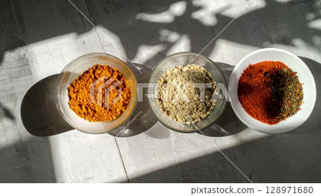 Overhead Shot of Turmeric Sesame and Chili Powder in Glass Bowls on Concrete Surface 128971680