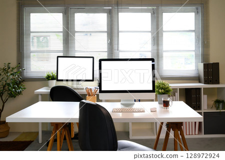 Home office interior with desktop computers on white desks and natural light from large windows 128972924