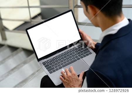 Male office worker sitting on stairs near a building, holding laptop with a white screen 128972926