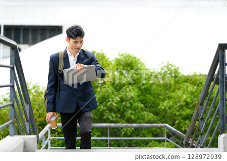 Businessman walking up stairs with laptop and coffee, looking at his watch 128972939