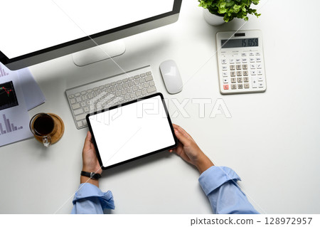 Overhead shot of hands holding blank screen digital tablet in front of a desktop computer Overhead shot of hands holding blank screen digital tablet in front of a desktop computer 128972957