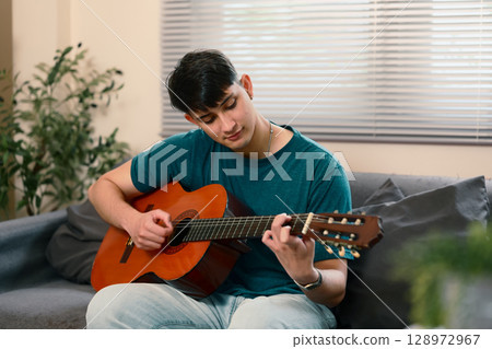 Young caucasian man sitting on a couch playing an acoustic guitar at home Young caucasian man sitting on a couch playing an acoustic guitar at home 128972967
