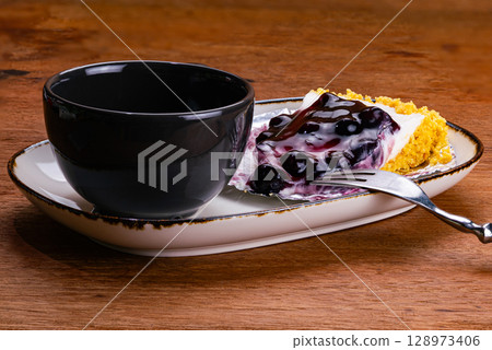 Side view of a cup of coffee in black ceramic cup with blueberry cheese pie on foil paper. 128973406