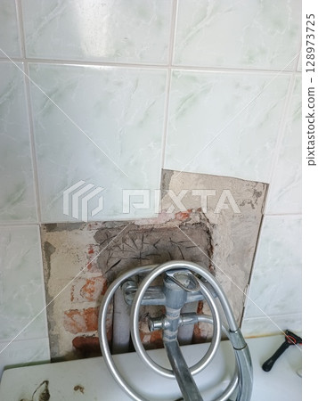 Damaged Kitchen Floor Tile Removal: Brick Substrate Exposed, Plumbing Fixture, Rust, and Disrepair in a Beige Tiled Interior Space 128973725