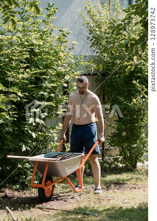 Shirtless Man Gardening Outdoors with Wheelbarrow, Lush Green Bushes, Summer Yard, Sunlight, Casual Scene, Orange Tool, Rustic Texture 128973742