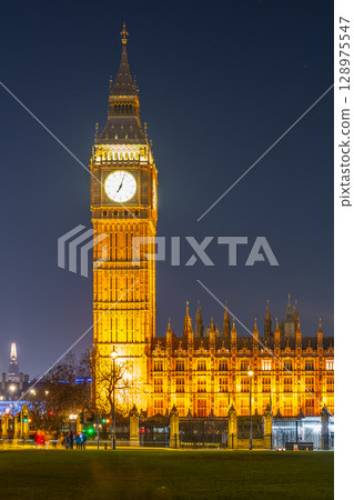 Big Ben stands proudly against the night sky, its clock face glowing brilliantly. The historic structure, surrounded by trees, showcases the beauty of London after sunset. 128975547