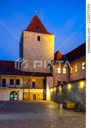 The Black Tower stands tall at Prague Castle, bathed in warm light against the night sky. The cobblestone courtyard glimmers under lamp light, inviting exploration. The Black Tower stands tall at Prague Castle, bathed in warm light against the night sky. The cobblestone courtyard glimmers under lamp light, inviting exploration. 128975549