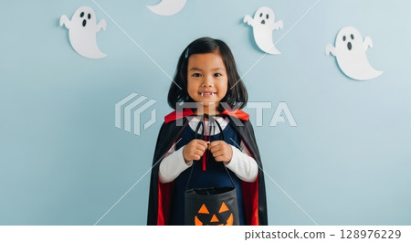 Cheerful asian girl in vampire costume with fangs and halloween candy bucket 128976229