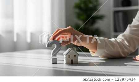 Woman hand placing question mark next to miniature house model on a desk 128976434
