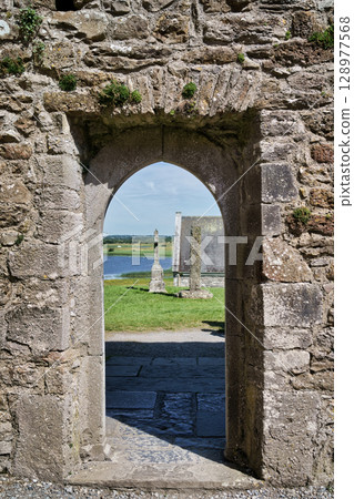 View through an archway of an Irish historical site, showcasing a tranquil landscape with a cross and church ruins. 128977568