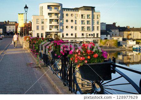 A scenic view in Ireland showcasing a vibrant display of flowers along a bridge, with buildings reflecting in the water. 128977629