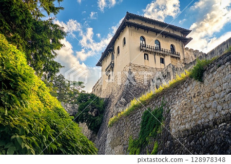 Castle with brightly shining walls stands cheerfully against a backdrop of majestic mountains and clear blue skies 128978348