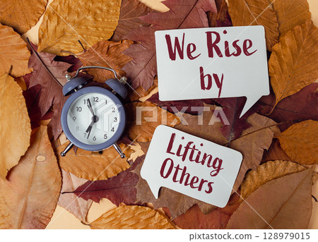 Clock is on a pile of leaves with the words "we rise by lifting others" writte 128979015