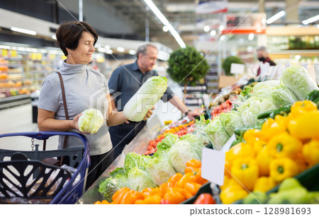 elderly woman examines china cabbage in vegetable section to choose juiciest cabbage elderly woman examines china cabbage in vegetable section to choose juiciest cabbage 128981693