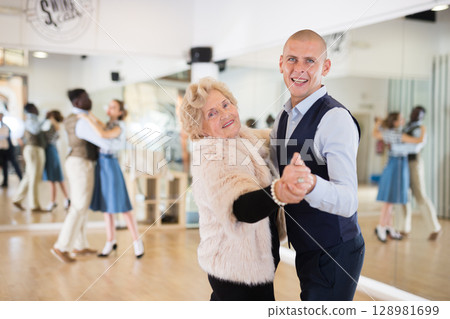 Man and elderly woman performing ballroom dance in dancing room Man and elderly woman performing ballroom dance in dancing room 128981699