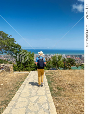A man wearing a hat and carrying a backpack walks along a stone path toward a panoramic view A man wearing a hat and carrying a backpack walks along a stone path toward a panoramic view 128982142