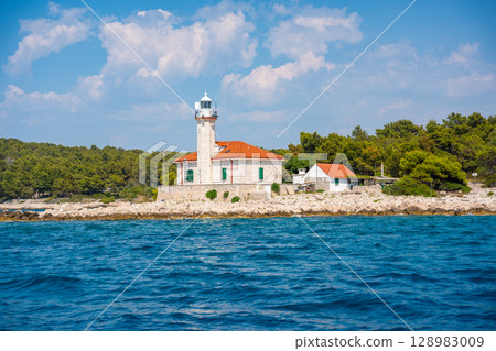Stone lighthouse with red roof on the rocky coast of Hvar Island, Croatia, surrounded by pine trees and clear blue sea. Navigation, solitude and maritime heritage in the Adriatic. 128983009