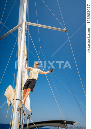 Man climbing the mast of a sailing yacht with full sails, captured from above near Croatia. Courage, freedom, yachting lifestyle and maritime exploration. Man climbing the mast of a sailing yacht with full sails, captured from above near Croatia. Courage, freedom, yachting lifestyle and maritime exploration. 128983017