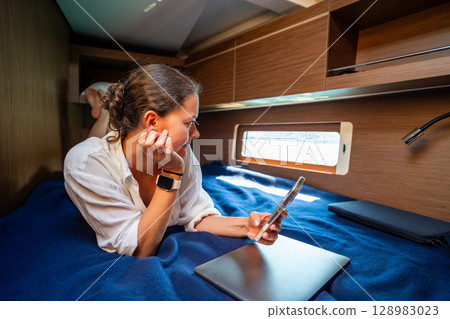 Young woman working with laptop and smartphone in the cabin of a sailing yacht near Croatia. Remote work, digital freedom and opportunity to live and work at sea. 128983023