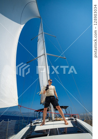 Man standing with smartphone on the bow of a sailing yacht under full sails near Croatia. Blogging, digital lifestyle, freedom and remote travel by sea. 128983034