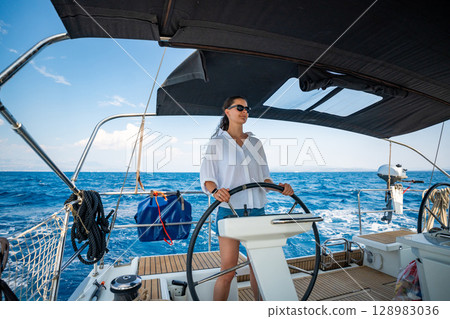 Young woman steering a sailing yacht near Croatia, confidently standing at the helm. Female leadership, empowerment and freedom in maritime lifestyle. 128983036