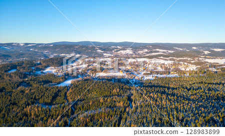 Panoramic view of Nova Ves nad Nisou showcasing the snow-covered landscape of the Jizera Mountains during a clear winter afternoon. Forests blanket the hills under a bright blue sky. Panoramic view of Nova Ves nad Nisou showcasing the snow-covered landscape of the Jizera Mountains during a clear winter afternoon. Forests blanket the hills under a bright blue sky. 128983899