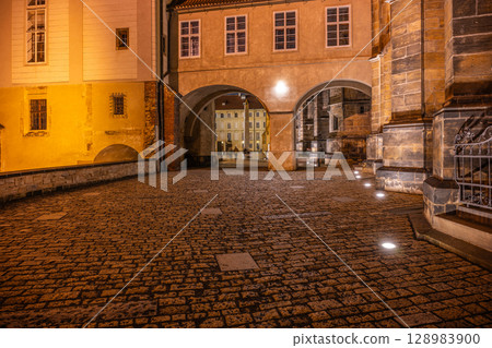The covered bridge at Saint Vitus Cathedral in Prague Castle glows softly under lamp light, creating a magical atmosphere at night. Visitors explore the cobblestone paths nearby. The covered bridge at Saint Vitus Cathedral in Prague Castle glows softly under lamp light, creating a magical atmosphere at night. Visitors explore the cobblestone paths nearby. 128983900
