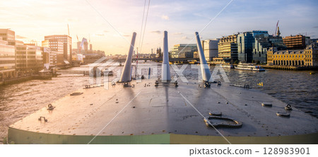 Turrets of HMS Belfast stand prominently along the River Thames, with the London skyline visible in the background during a peaceful sunset. 128983901