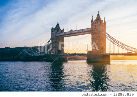 Tower Bridge stands majestically at sunrise, with warm hues illuminating its structure. The calm waters reflect the vibrant colors, showcasing a tranquil moment in the heart of London. 128983939
