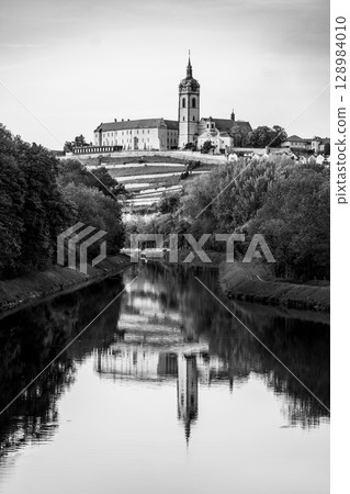 Chateau Melnik stands majestically on a hilltop, overlooking a tranquil river. The historic architecture is perfectly mirrored in the still water, showcasing its beauty in black and white. Chateau Melnik stands majestically on a hilltop, overlooking a tranquil river. The historic architecture is perfectly mirrored in the still water, showcasing its beauty in black and white. 128984010
