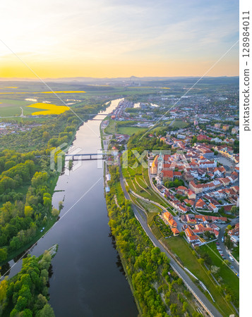 An aerial view captures Melnik City with its historic chateau perched above the serene Labe River, surrounded by green landscapes and rolling hills during golden hour. 128984011