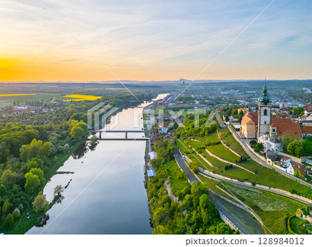 Aerial view captures Melnik city and its historic chateau perched above the tranquil Labe River. The golden hour illuminates the landscape, showcasing lush greenery and flowing waters. 128984012