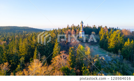 On a sunny spring morning, the Stepanka Lookout Tower stands majestically atop green hills in Korenov, surrounded by lush forests and a clear blue sky. 128984016